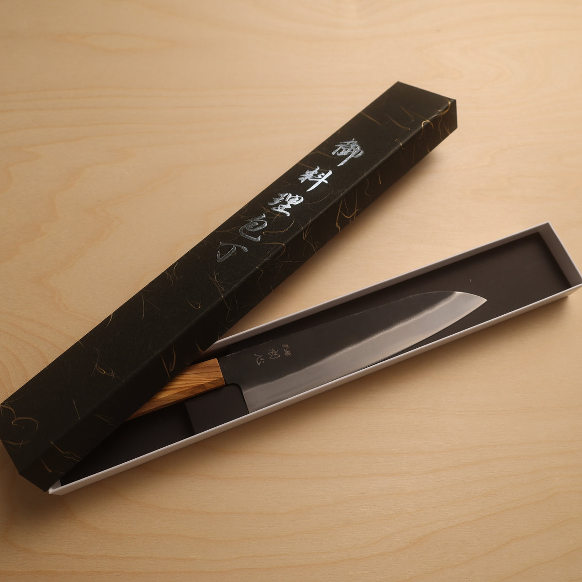 A Japanese kitchen knife with a wooden handle and silver blade, presented in an open black gift box with Japanese characters on the lid, set on a light wood background.