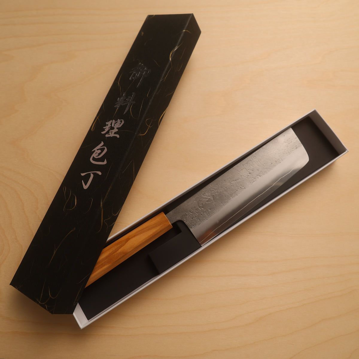 A Japanese-style kitchen knife with a patterned steel blade and a light wood handle, presented in an open black box with gold calligraphy on a light wood tabletop.