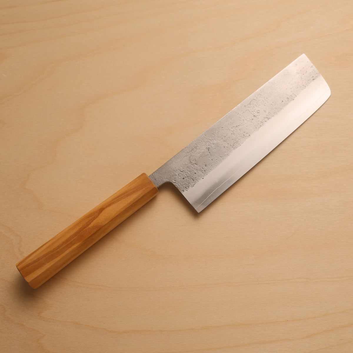 A Japanese-style kitchen knife with a textured, patterned steel blade and a light brown wooden handle, placed diagonally on a light wood grain background.