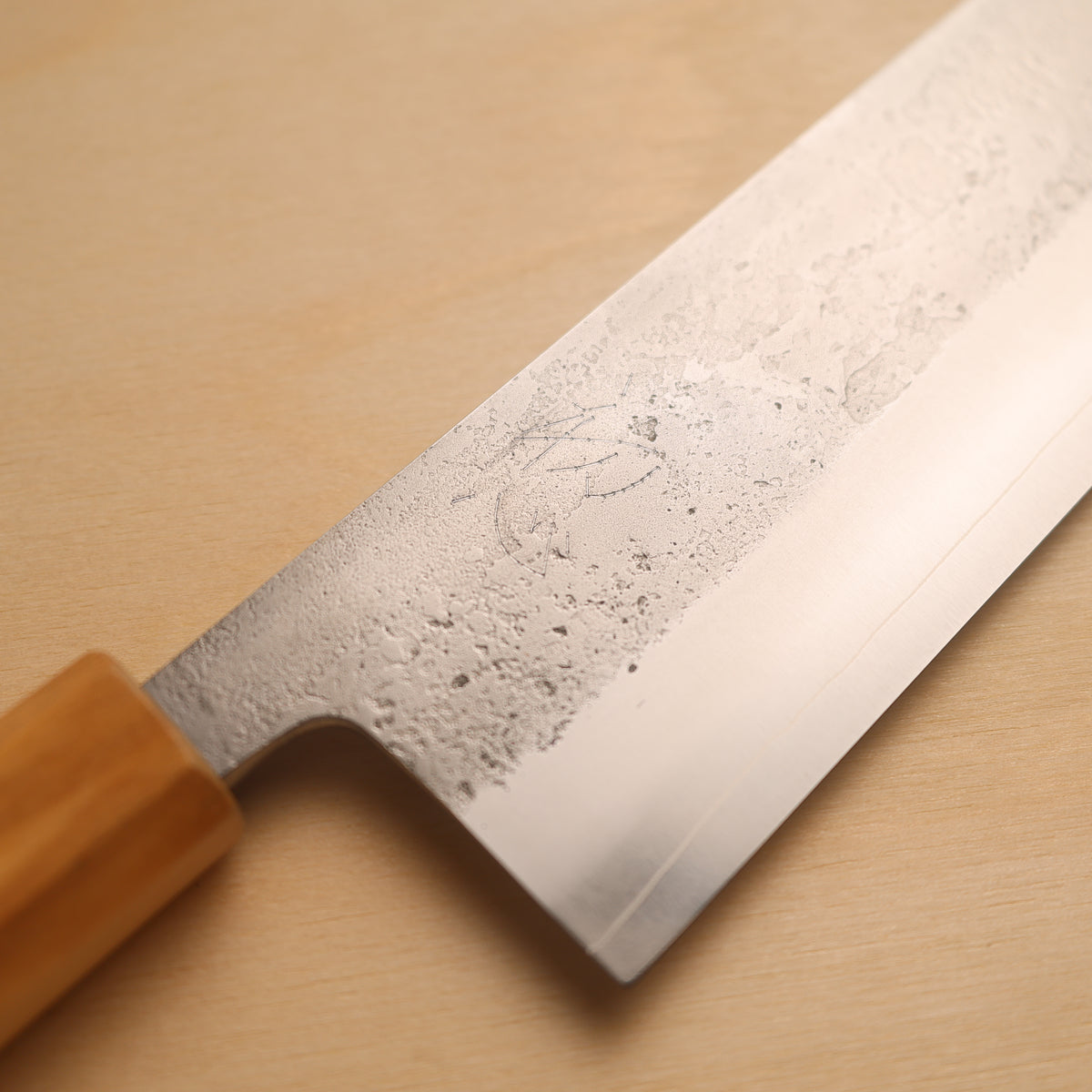A close-up of a Japanese-style kitchen knife with a textured, hammered steel blade and a light brown wooden handle, resting diagonally on a light wood grain surface.