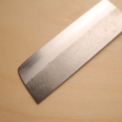 A close-up of a Japanese-style kitchen knife blade with a textured, hammered finish, resting diagonally on a light wood grain surface.
