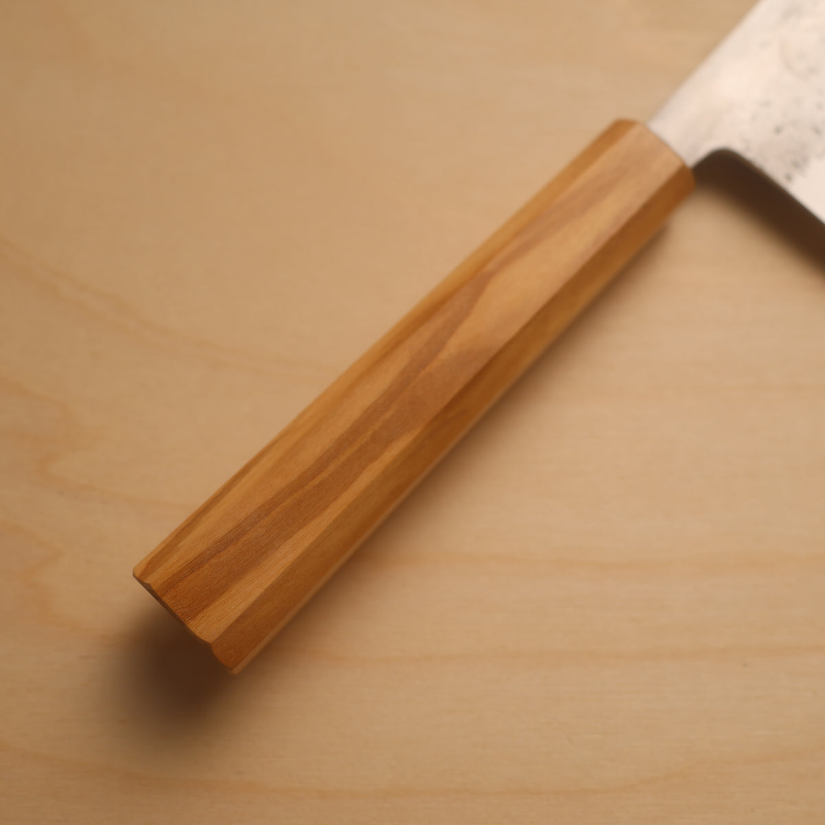 A close-up of a Japanese-style kitchen knife with a hexagonal wooden handle and a textured steel blade, resting on a light wood surface.