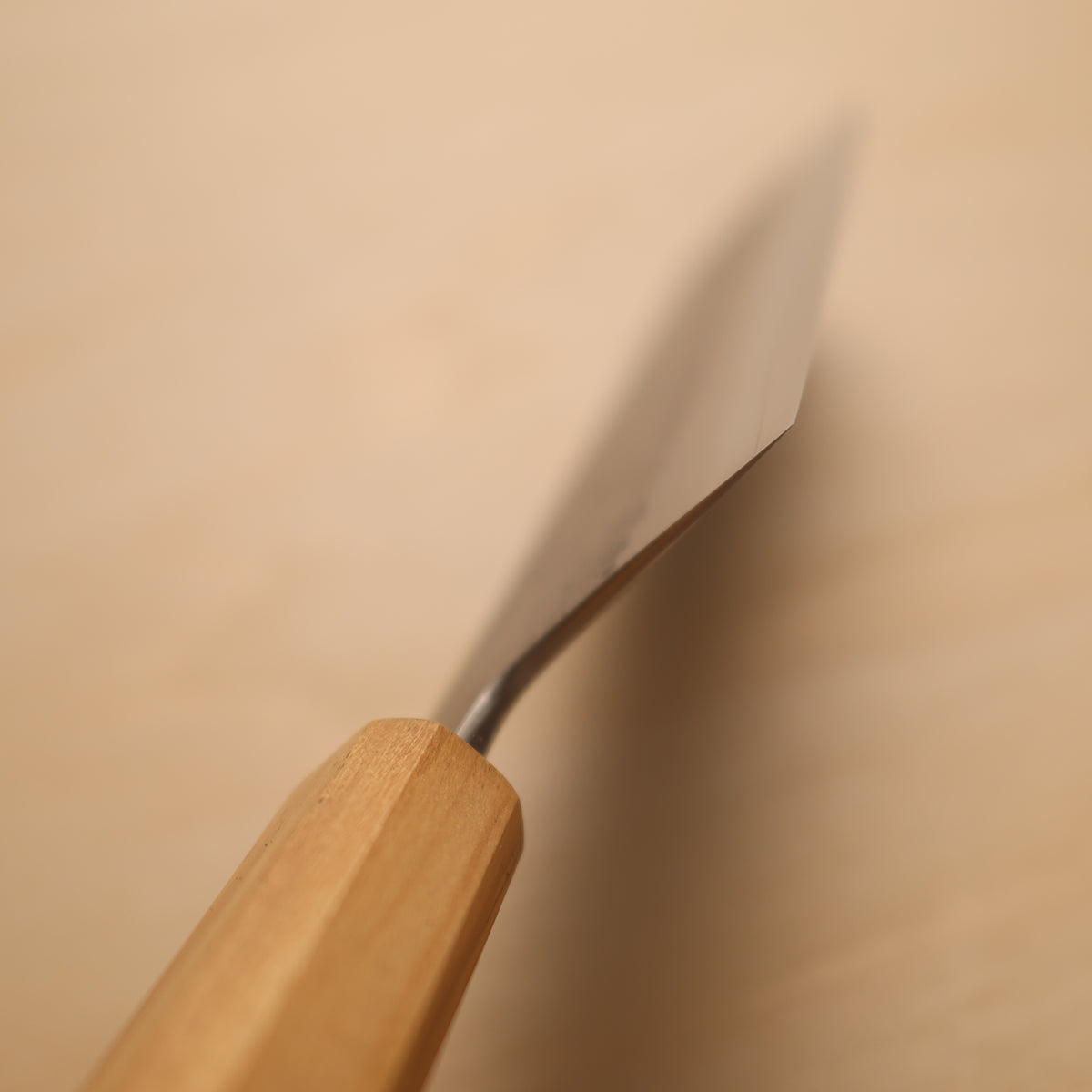 A close-up of a hand tool with a light natural wood handle and a silver metal blade, shown against a warm beige background.