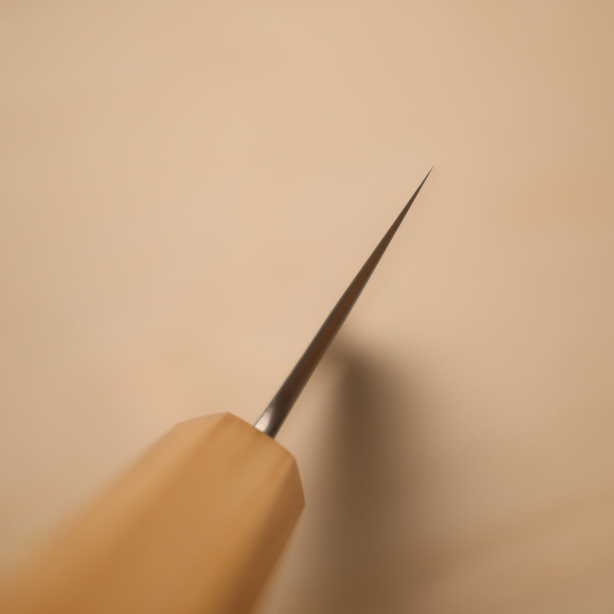 A close-up of a hand tool with a light natural wood handle and a very sharp metallic needle-like tip, photographed against a warm beige background.