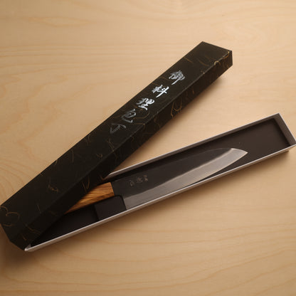 A Japanese kitchen knife with a wooden handle and silver blade, presented in an open black gift box with Japanese characters on the lid, set on a light wood background.