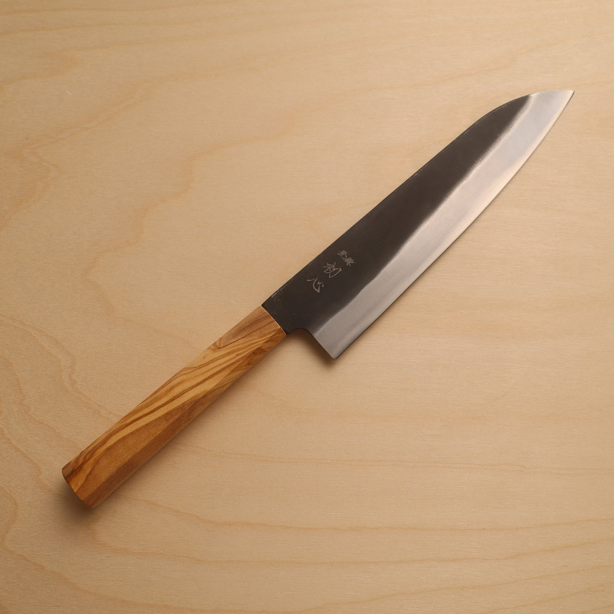 A Japanese-style kitchen knife with a polished steel blade and a natural olive wood handle, placed diagonally on a light wood grain surface.