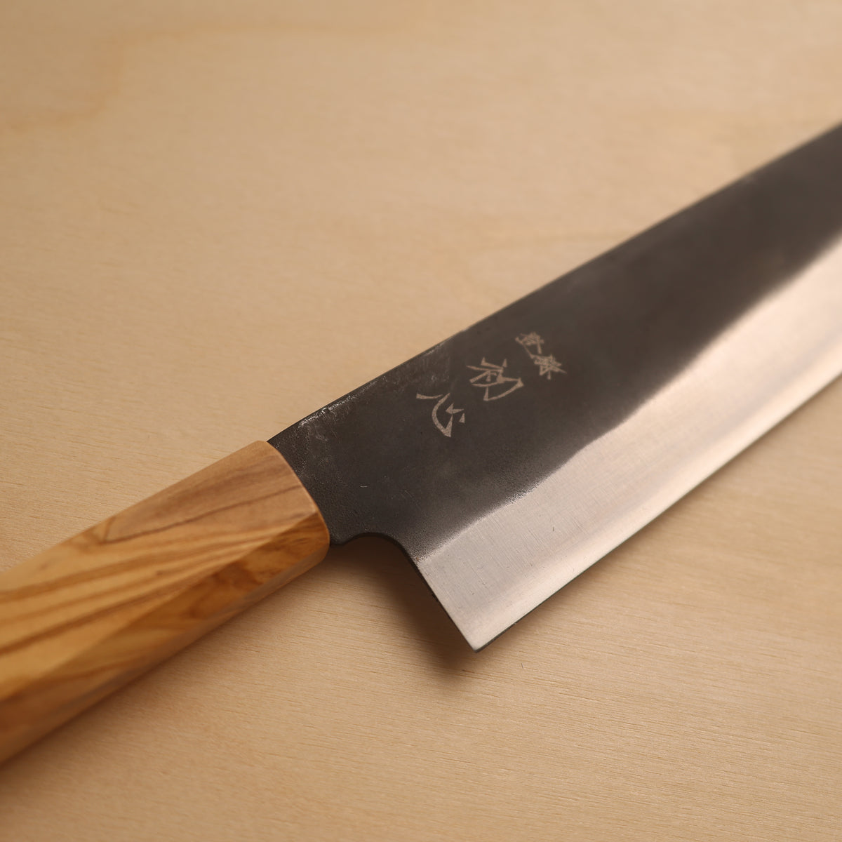 A Japanese-style kitchen knife with a polished steel blade featuring engraved kanji characters and a light brown wooden handle, resting diagonally on a light beige wooden surface.