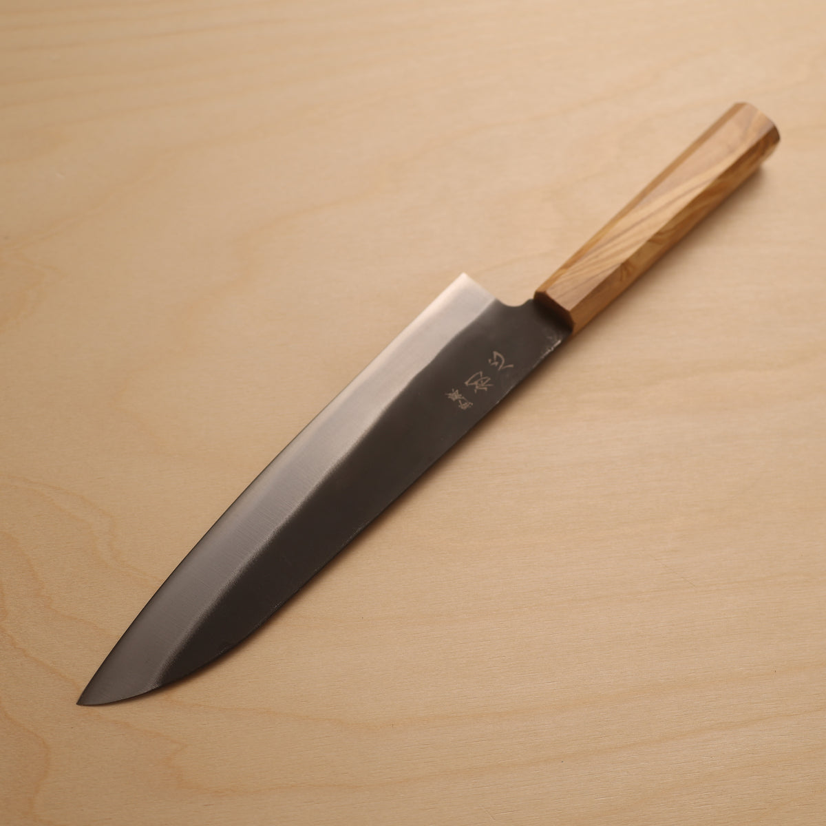 A Japanese kitchen knife with a dark steel blade and a light wood handle, placed diagonally on a light wood grain surface.