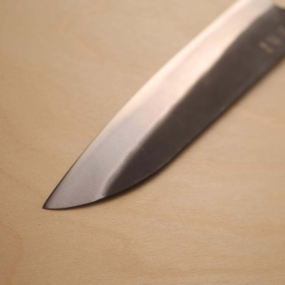 A close-up of a Japanese-style kitchen knife blade resting diagonally on a light-colored wooden surface.