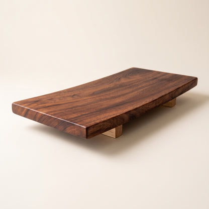 A rectangular live-edge wooden serving board or low coffee table with a rich dark brown wood grain, supported by two small wooden blocks on a warm beige background.
