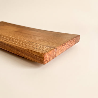 A close-up of a rectangular wooden board with rounded corners, showing a warm medium-brown wood grain and a lighter, slightly pinkish end grain, photographed on a clean off-white background.