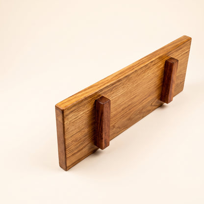 A rectangular wooden board with a warm honey-toned wood grain finish and two darker reddish-brown wooden pegs or supports, photographed on a clean off-white background.