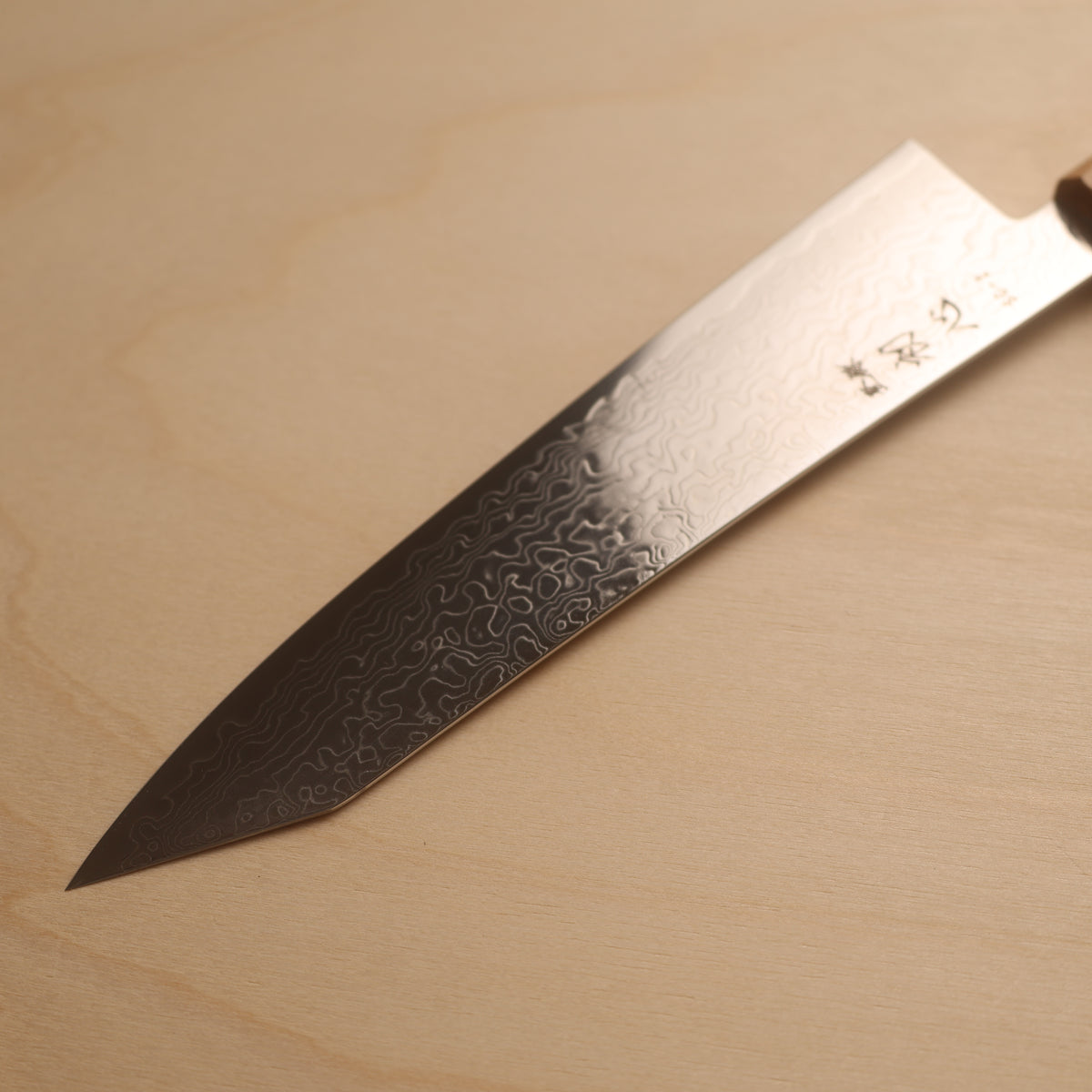 A Japanese kitchen knife with a Damascus steel blade, featuring a wavy pattern and engraved characters, resting diagonally on a light wood background.