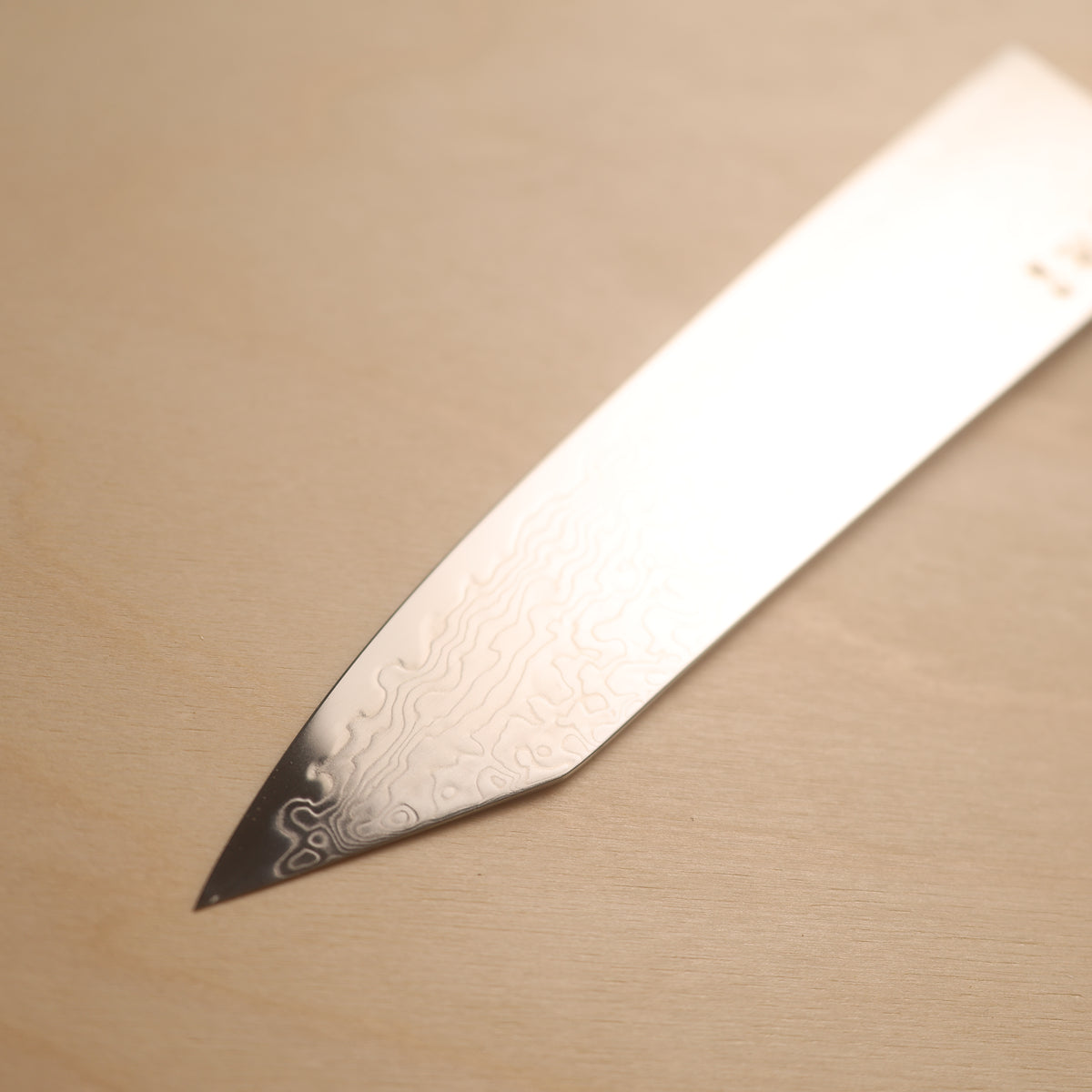 A close-up of a Japanese-style chef's knife tip with a Damascus steel pattern, resting on a light beige wooden surface.