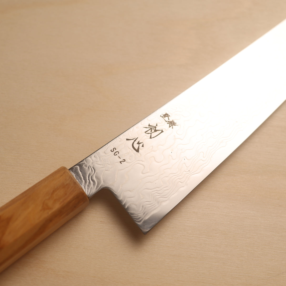 A Japanese kitchen knife with a Damascus steel blade and a light brown wooden handle, shown diagonally on a light wood background. The blade features engraved kanji characters and the model name 'SG-2'.