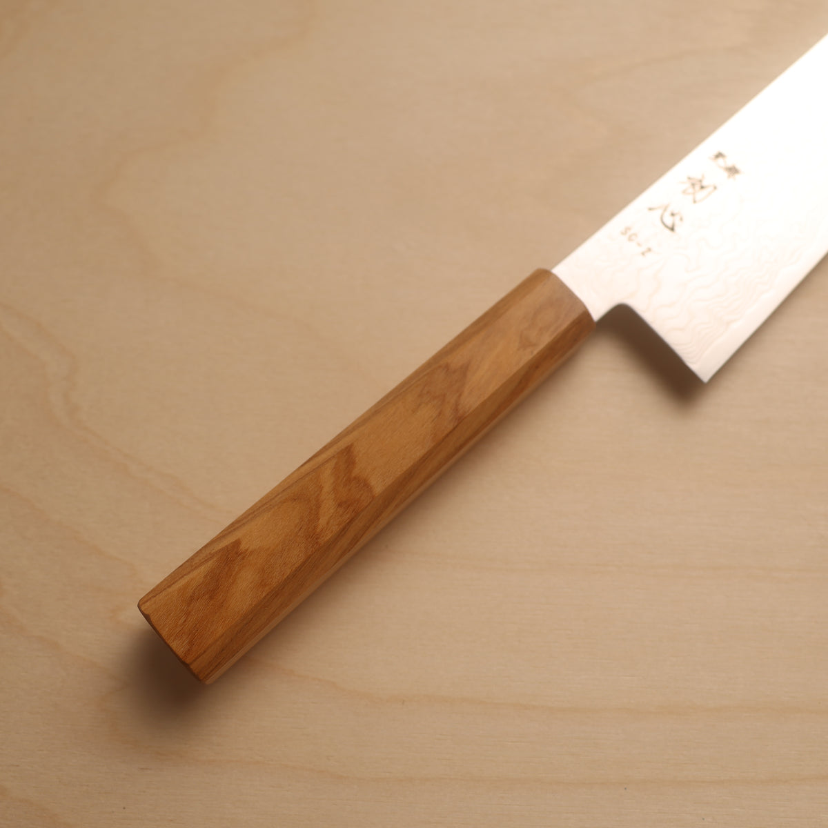 A Japanese-style kitchen knife with a light brown wooden handle and a silver steel blade, placed diagonally on a light wood grain surface. The blade features engraved Japanese characters.