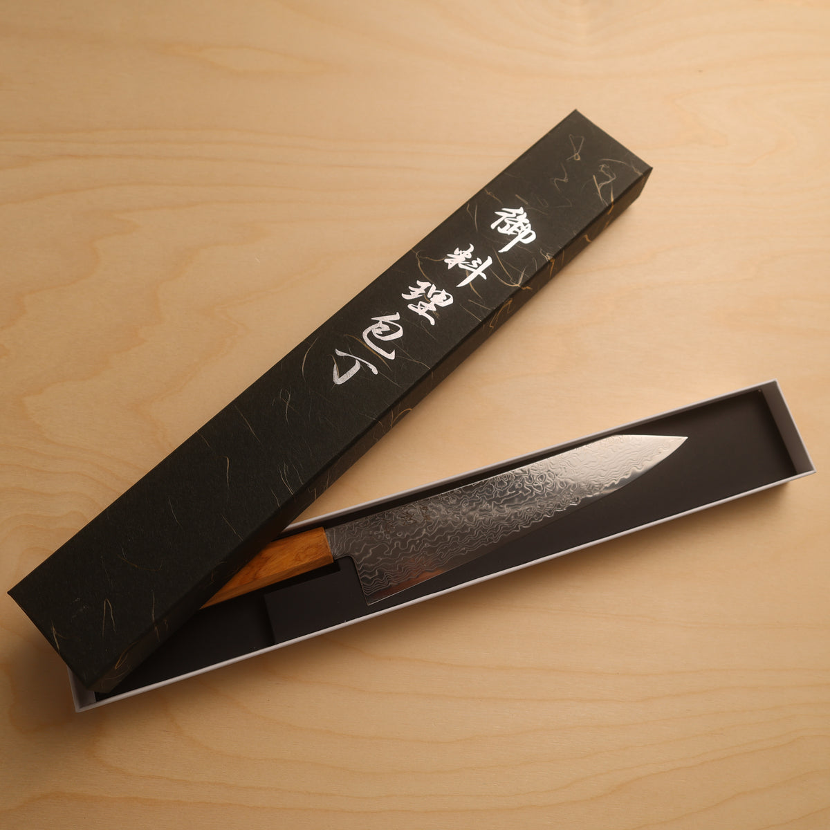 A Japanese-style kitchen knife with a patterned steel blade and wooden handle, partially pulled from its black presentation box on a light wood tabletop. The box has white Japanese calligraphy.