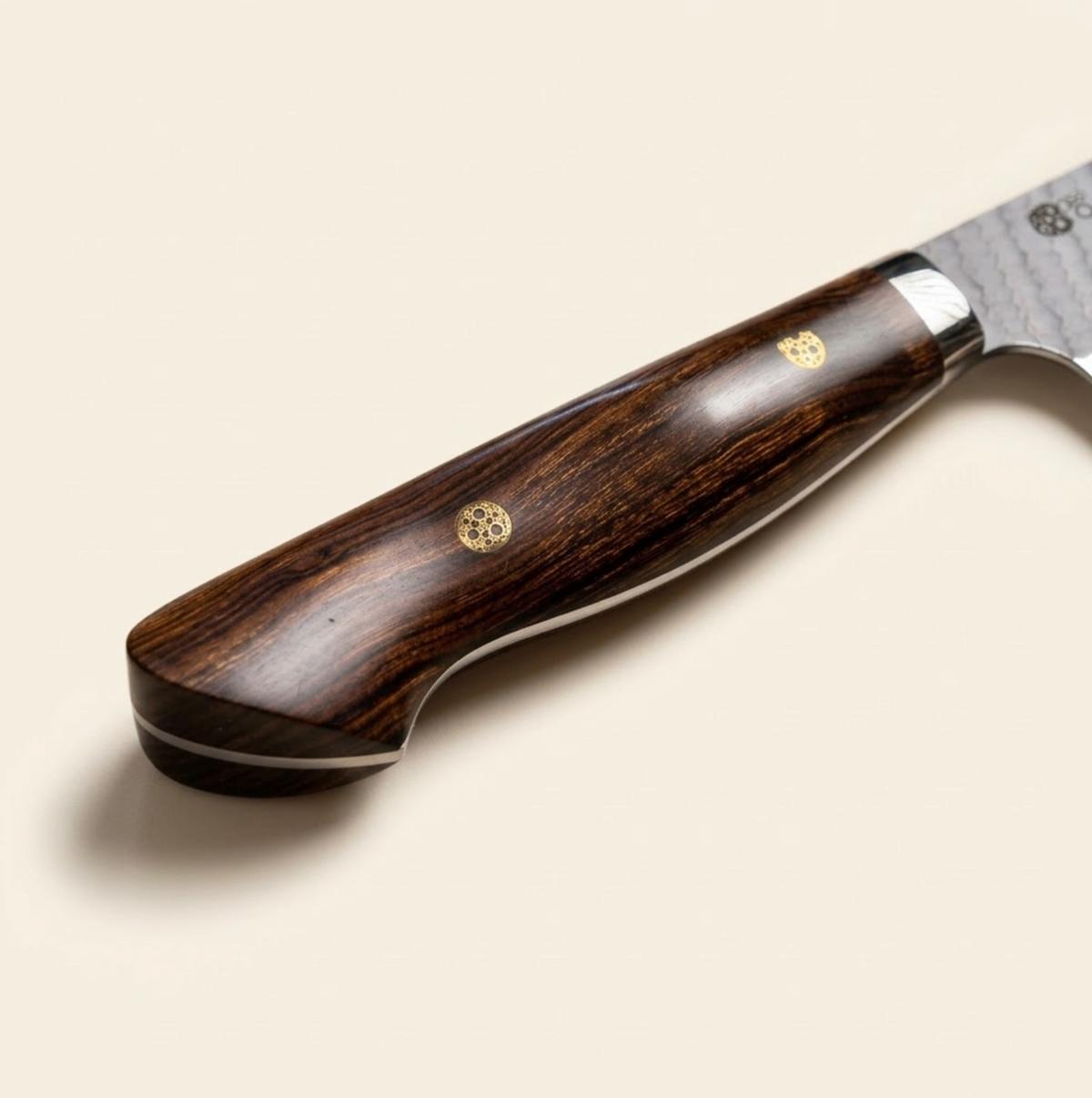 nigara A close-up of a high-end kitchen knife handle made of dark brown wood with two gold-toned circular inlays, set against a solid light beige background. iron wood handle