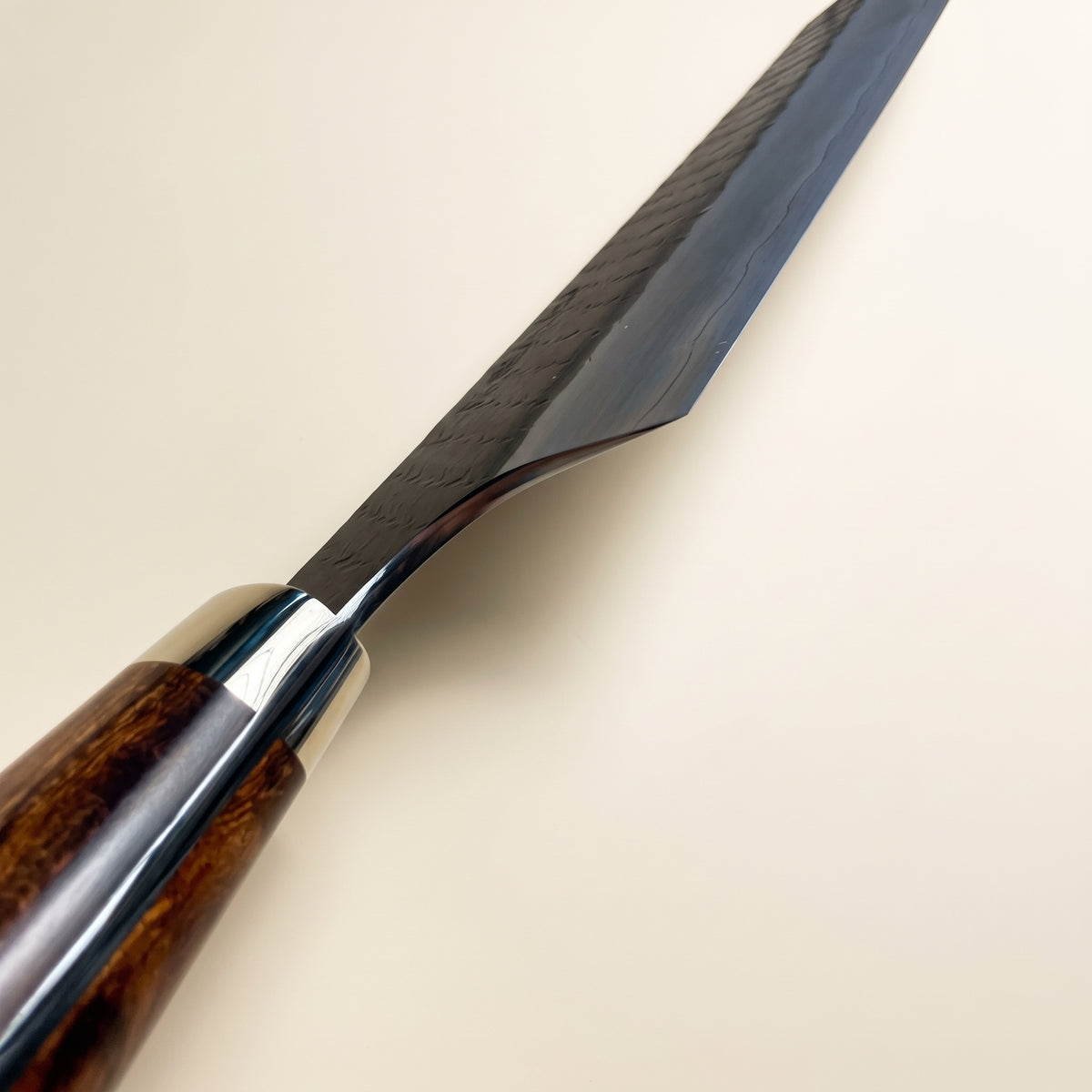A close-up of a Japanese-style kitchen knife with a dark patterned steel blade and a polished dark wood handle, shown against a solid light beige background.
