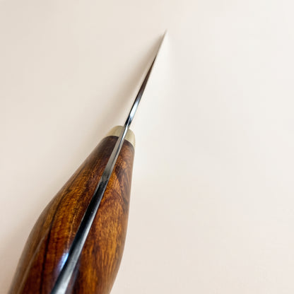 A close-up of a hand tool with a polished dark brown wooden handle and a sharp silver metal edge, positioned diagonally against a plain off-white background. Nigara knife choil polish with western handle
