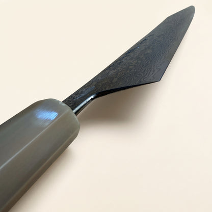 A close-up of a tojiro atelier kitchen knife with a dark patterned Damascus-style blade and a smooth gray handle, set against a plain light beige background.