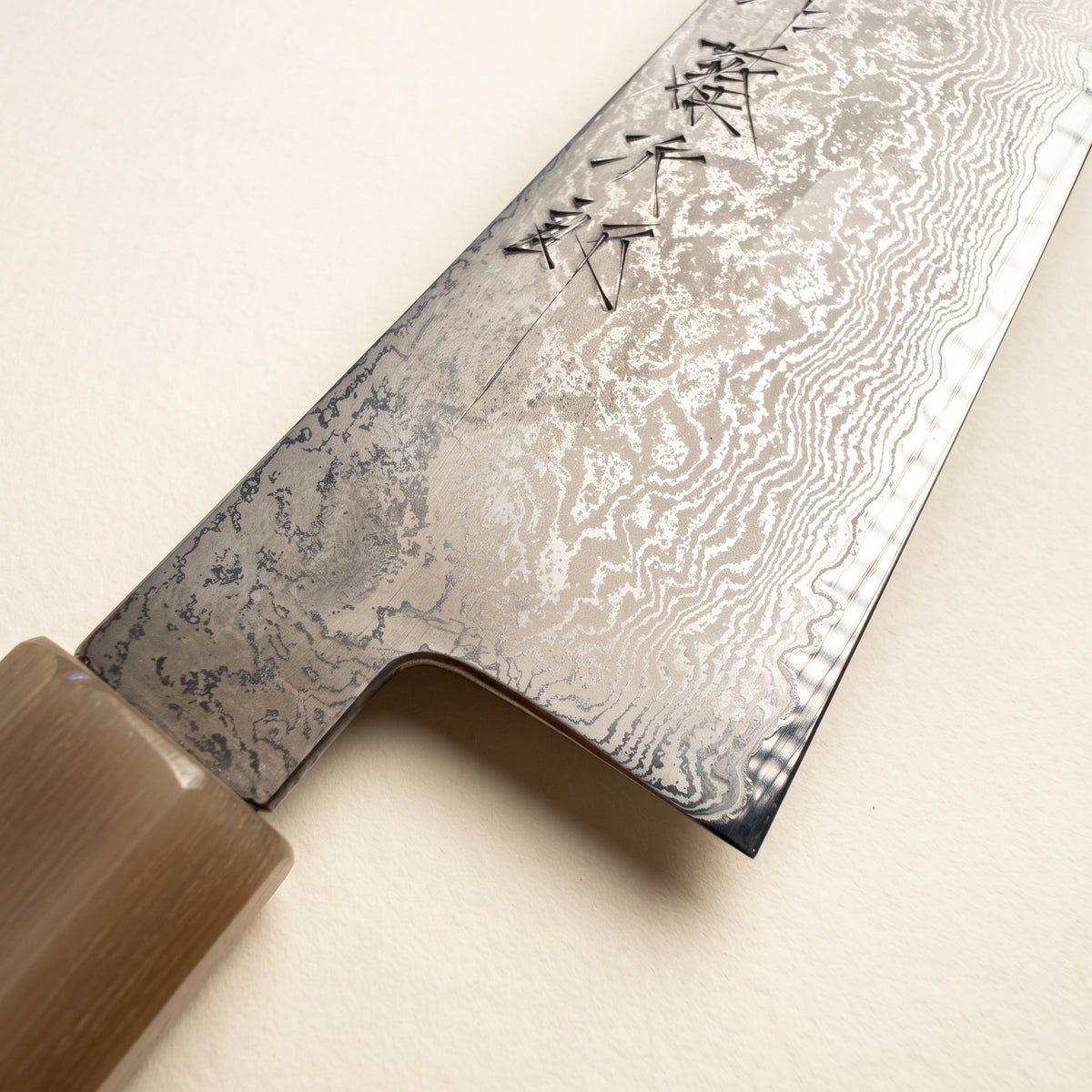 A close-up of a Japanese kitchen knife showing its Damascus steel blade with black kanji characters engraved near the tip, and a light brown wooden handle, set against a plain off-white background.